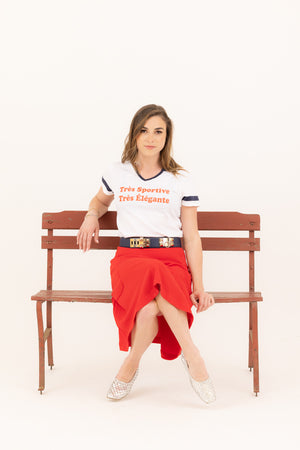 Young woman sitting on a bench wearing a graphic baseball tee that says "trés sportive, trés èlègante paired with a red Mireille midi skirt.