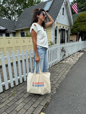 Woman in the white Julie top and ripped jeans holding a "le weekend" tote bag, standing in front of a charming house with a picket fence.