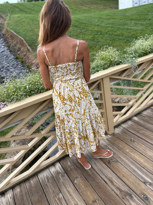 Woman in a strappy floral dress standing with her back towards us  on a wooden bridge over a stream with greenery in the background