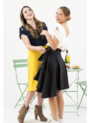 Two friends meeting at a cafe, one wearing Valerie wrap skirt in grey and white pinstripe and Julie V back top in white and the other wearing Mireille yellow midi skirt and navy Julie V neck top from Le Mireille Marche.