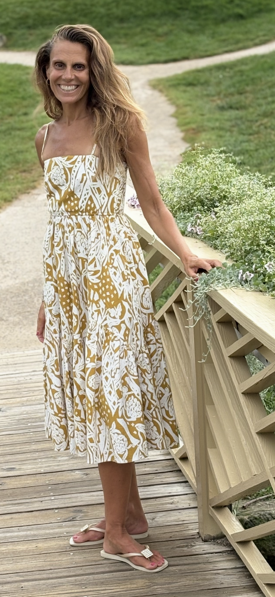Woman in a floral strappy dress with pockets standing on a wooden bridge with greenery in the background