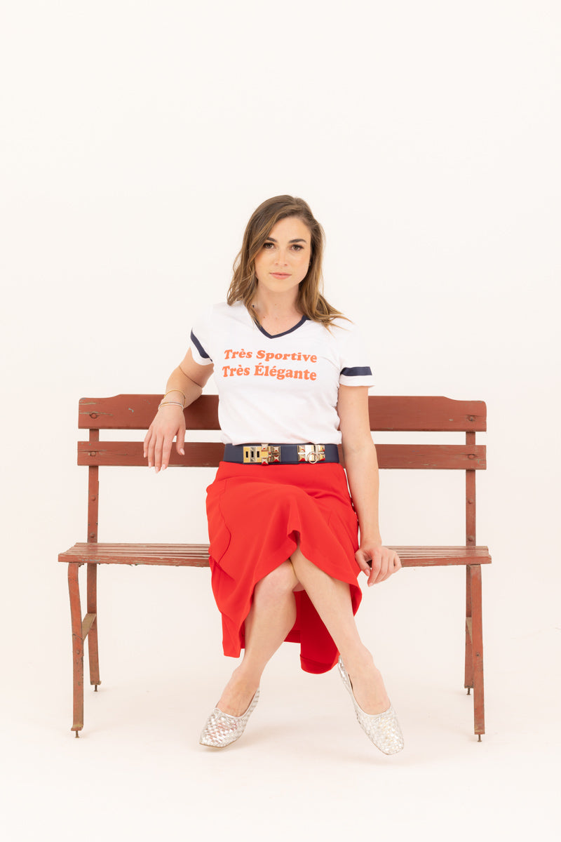 Young woman sitting on a bench wearing a graphic baseball tee that says &quot;trés sportive, trés èlègante paired with a red Mireille midi skirt.