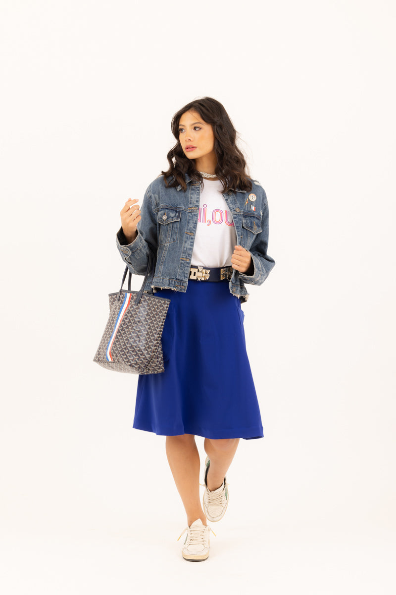 Raven haired woman wearing the denim Jean jacket, "oui, oui" graphic t-shirt, and electric blue midi skirt from Le Mireille Marche, accessorized with a Godard tote bag and sneakers.