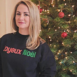 Blonde woman standing in front of her Christmas Tree wearing a top that says Merry Christmas in French.