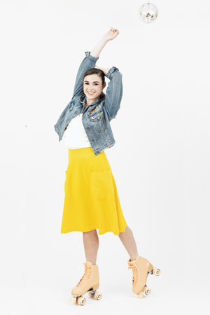 Woman on roller skates at a disco wearing denim Jean jacket with enamel pins, sporty Mireille midi skirt in yellow, and Julie top in white from Le Mireille Marche.