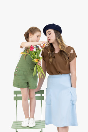 Woman in a brown Julie top and baby blue Midi skirt from Le Mireille Marche, wearing a beret, standing next to a child in green overalls holding tulips.