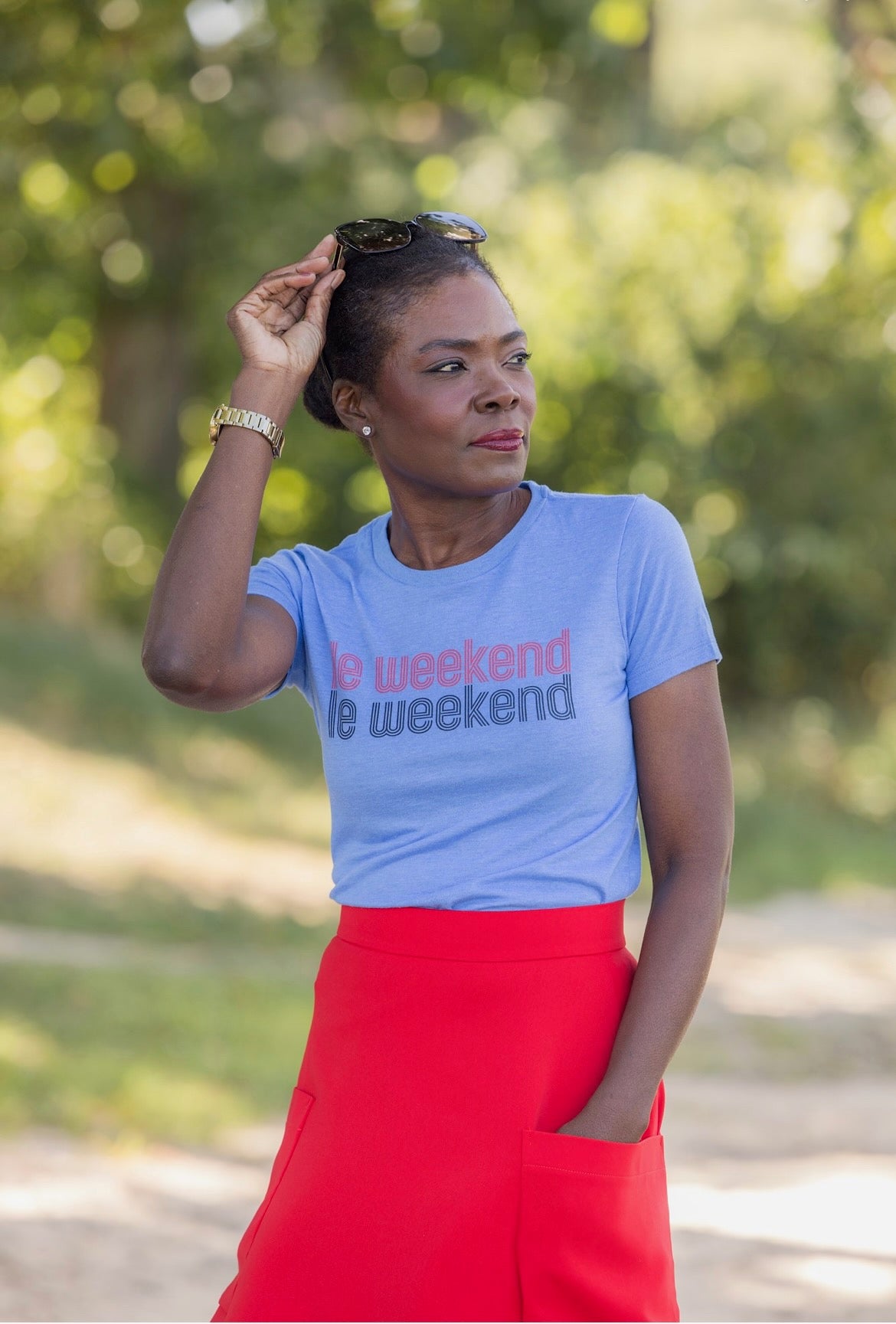 Chic woman standing in a park in a blue graphic tee shirt that says &quot;le weekend&quot; and a red midi skirt by Le Mireille Marche.