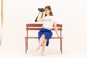 Casually chic woman sitting on a bench with a camera wearing an electric blue midi skirt and graphic tee shirt with the words "le weekend" from Le Mireille Marche.