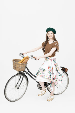 A French woman on her bicycle with a basket filled with bagettes wearing the Valerie skirt in the cherry blossom print and the Julie top in brown.