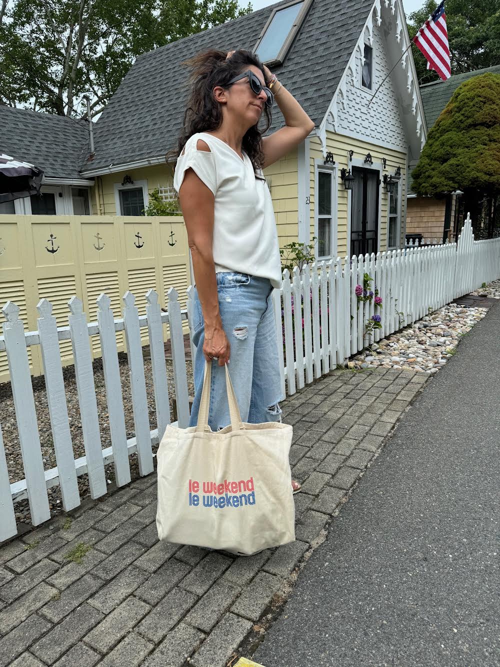 Woman in the white Julie top and ripped jeans holding a "le weekend" tote bag, standing in front of a charming house with a picket fence.