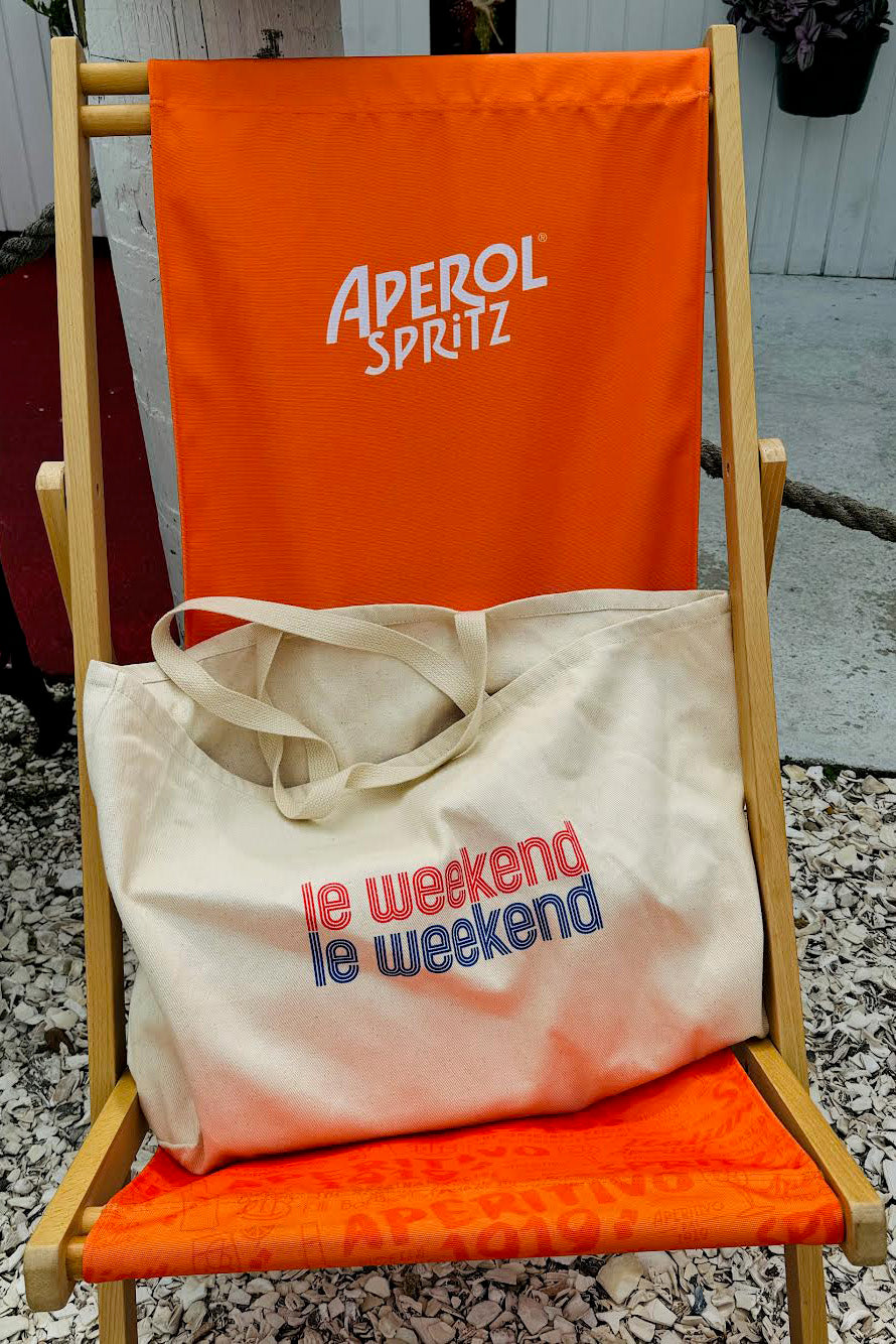 Canvas tote with the graphic "le weekend" in blue and red, on a canvas and wood outdoor chair.