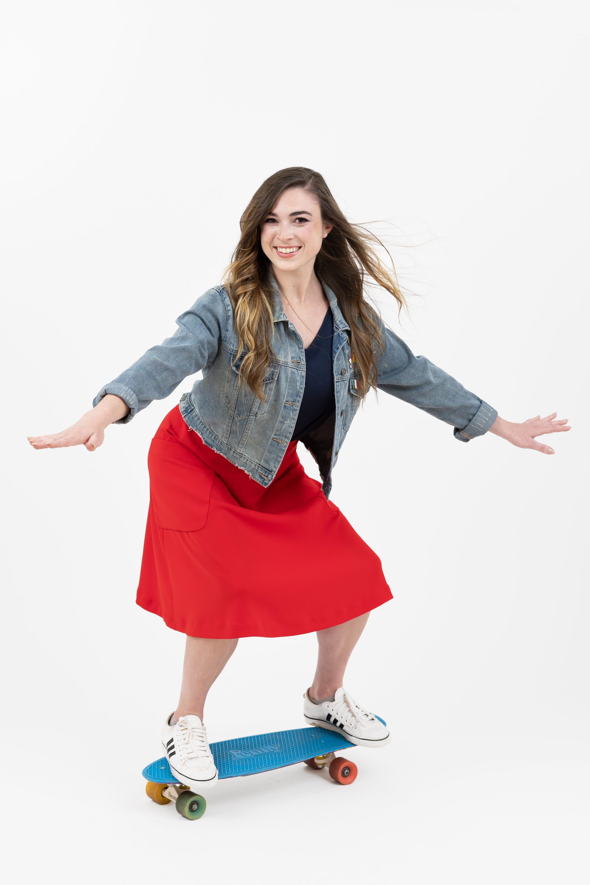 Woman skateboarding in a denim jacket and red skirt, smiling and balancing on a blue skateboard.