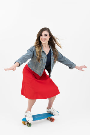 Athletic and elegant woman riding a skateboard wearing jean jacket and rouge/red Mireille two pocket midi skirt from Le Mireille Marche.