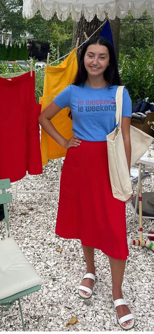 Woman wearing a blue "le weekend" t-shirt and red midi skirt from Le Mireille Marche, standing in an outdoor setting with colorful clothes hanging in the background.