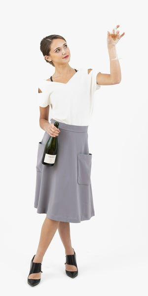 Young French woman wearing Julie top in blanche/white and Mireille midi skirt in grey celebrating with a bottle of champagne.